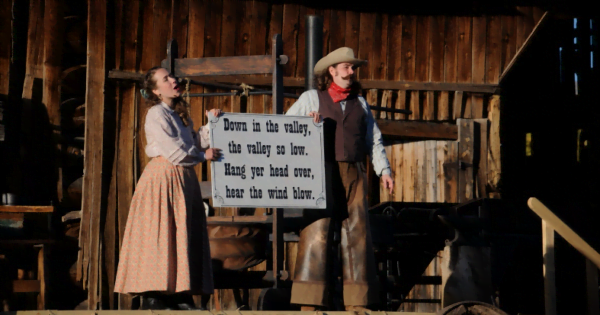 Photo of two performers in Western style setting and dress, holding a sign with song lyrics: "Down in the valley,/ the valley so low./ Hang your head over,/ hear the wind blow."